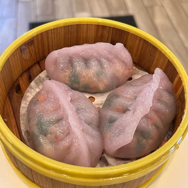 Three translucent pink dumplings with green and orange filling, in a bamboo steamer basket.