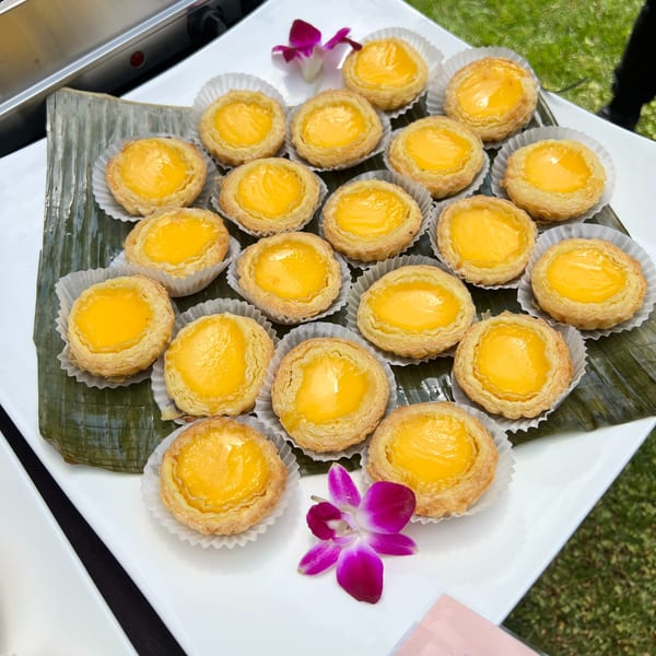 A white platter holds numerous golden-yellow egg tarts on banana leaves, garnished with purple orchids.
