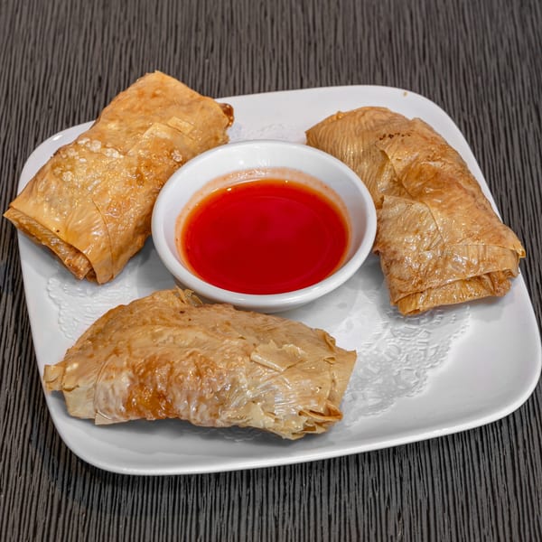 Four golden-brown fried bean curd skin rolls with a small bowl of red dipping sauce on a white plate.