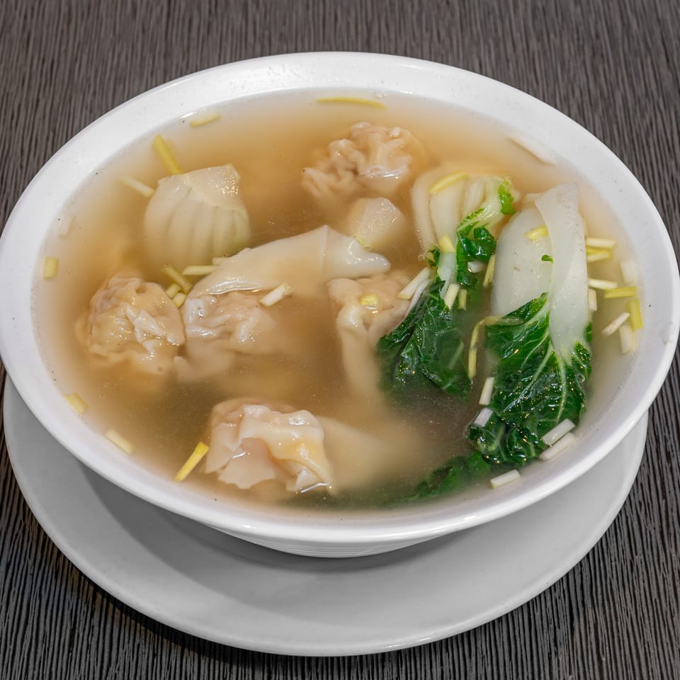 A bowl of Imperial Garden wonton soup with bok choy and scallions on a dark table.